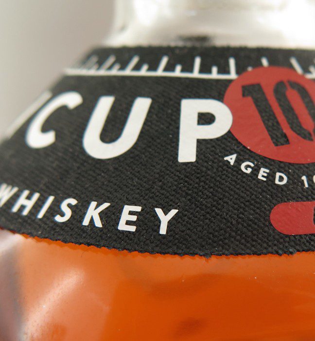 Close up photograph of a tin cup whiskey bottle label, showcasing Cpg packaging production expertise through distinctive on pack embellishments.