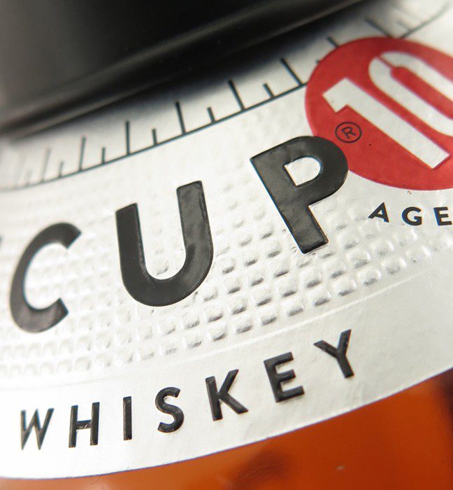 Close up photograph of a tin cup whiskey bottle label, showcasing Cpg packaging production expertise through distinctive on pack embellishments.